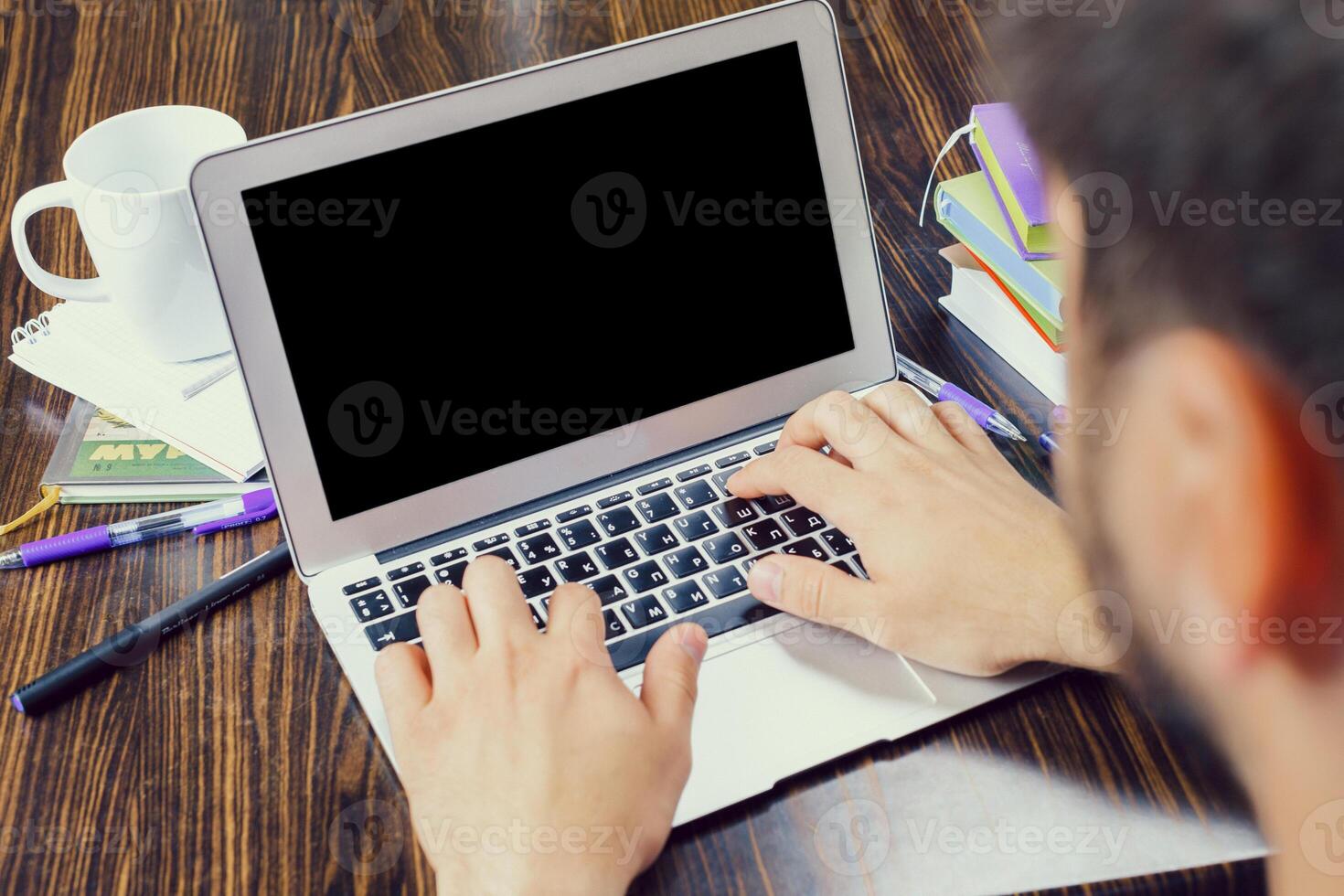 Person using laptop at a wooden table surrounded by school supplies in a study space during daytime photo