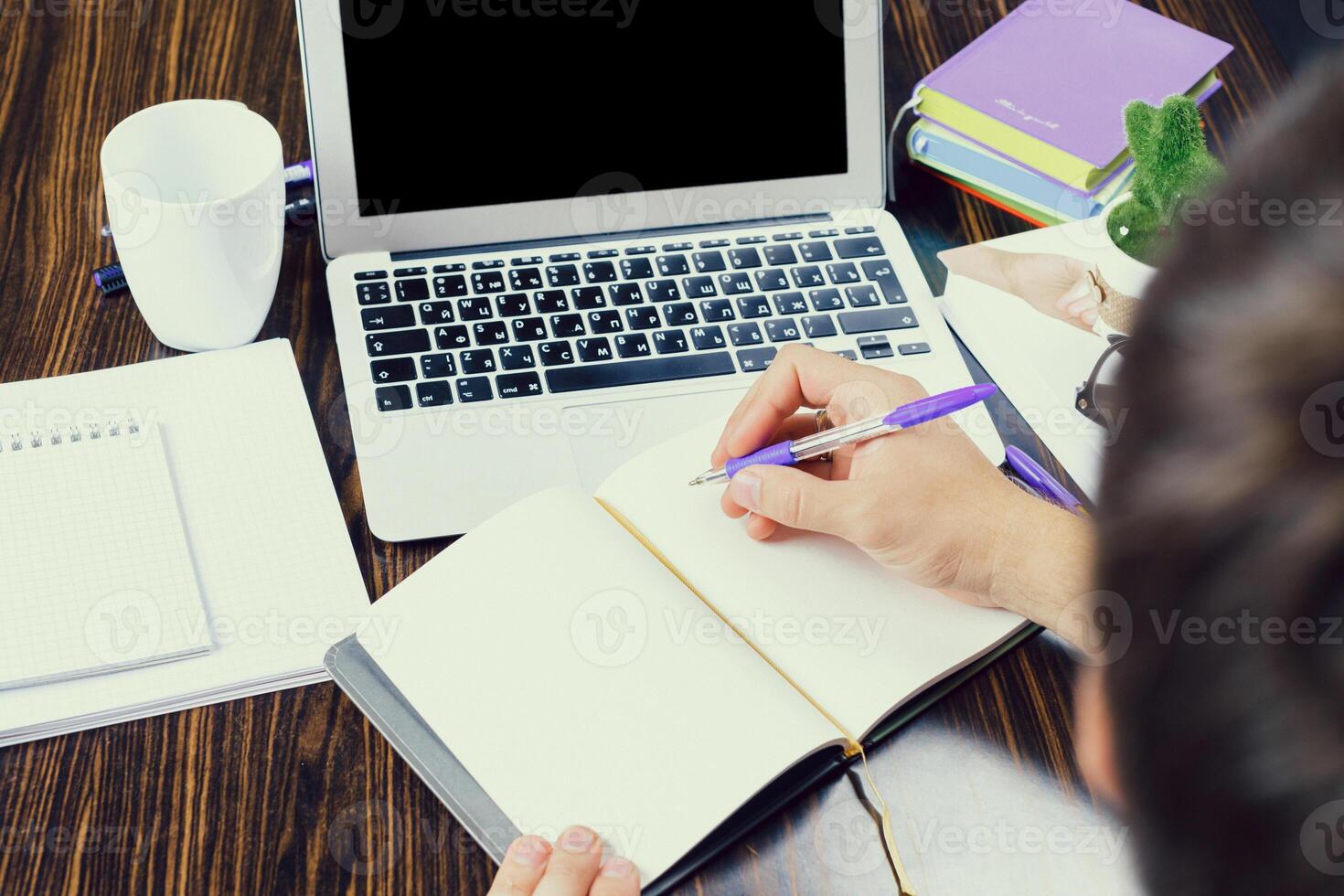 Person writing notes in a notebook beside a laptop and office supplies at a wooden desk during the day photo