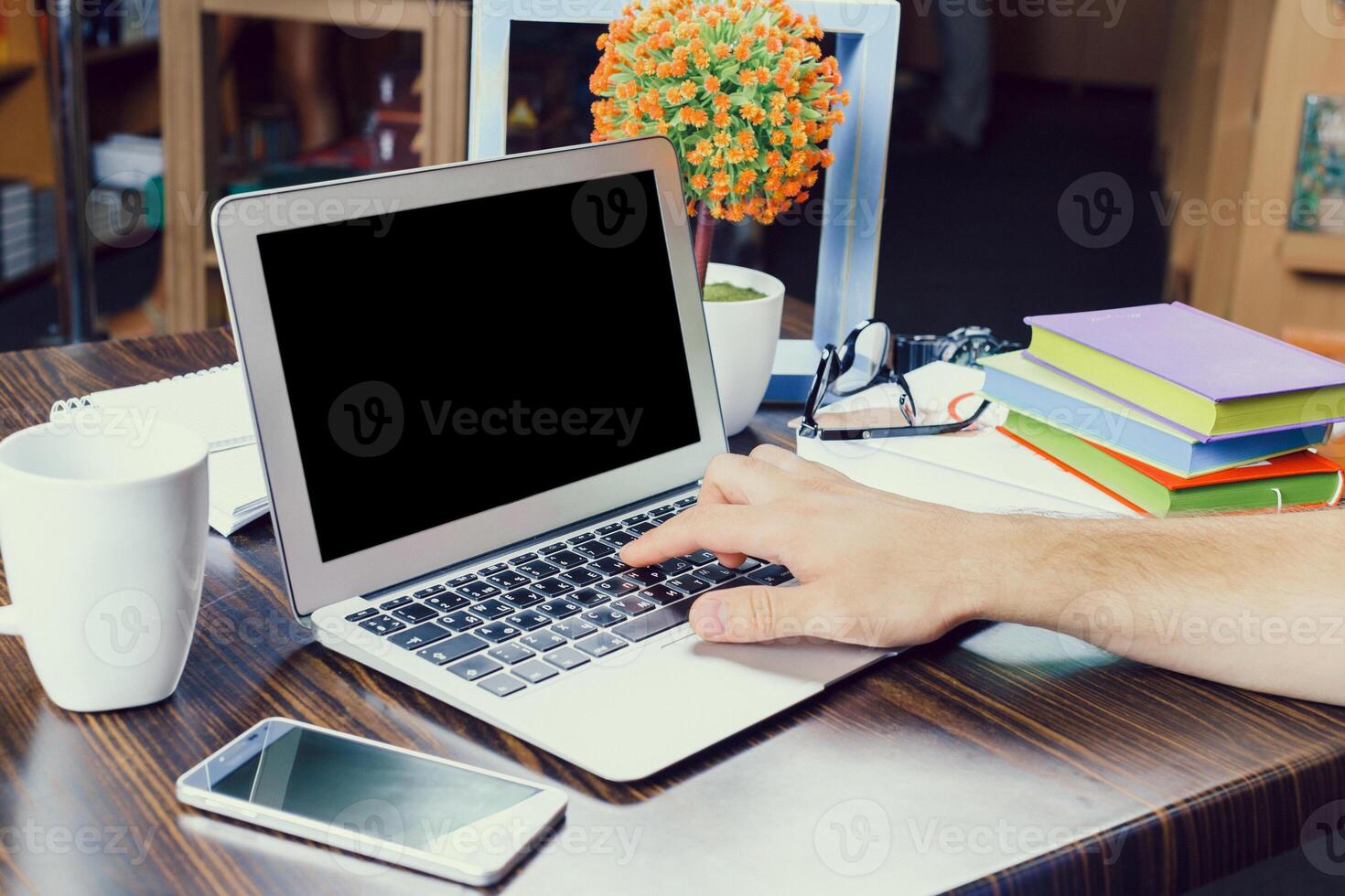 Person working on a laptop at a desk surrounded by books and plants in a cozy indoor setting photo