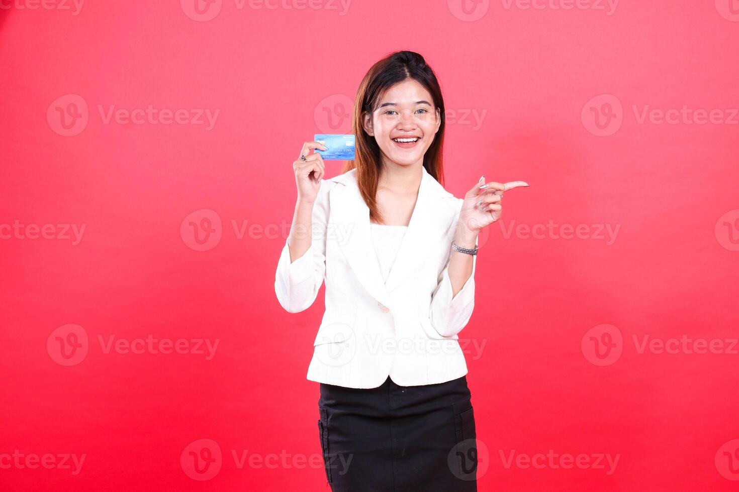 A woman in a white blazer and black skirt is holding and presenting a blue card, and pointing to the right, isolated on a red background photo
