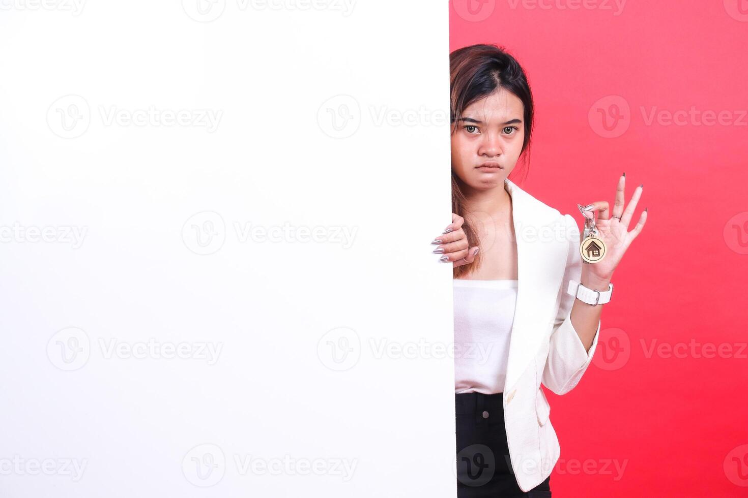 A woman partially visible behind a white board is holding keys with a house symbol on them, isolated on a red background. photo