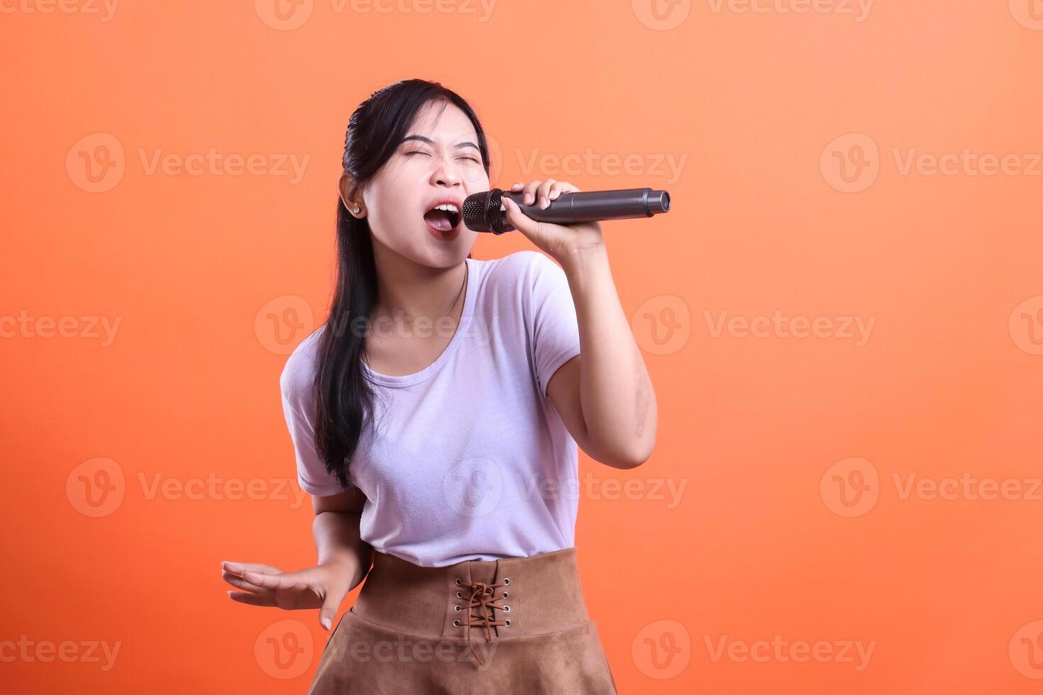 An enthusiastic young woman performs a song with a microphone, expressing pure joy and energy, isolated on orange background photo