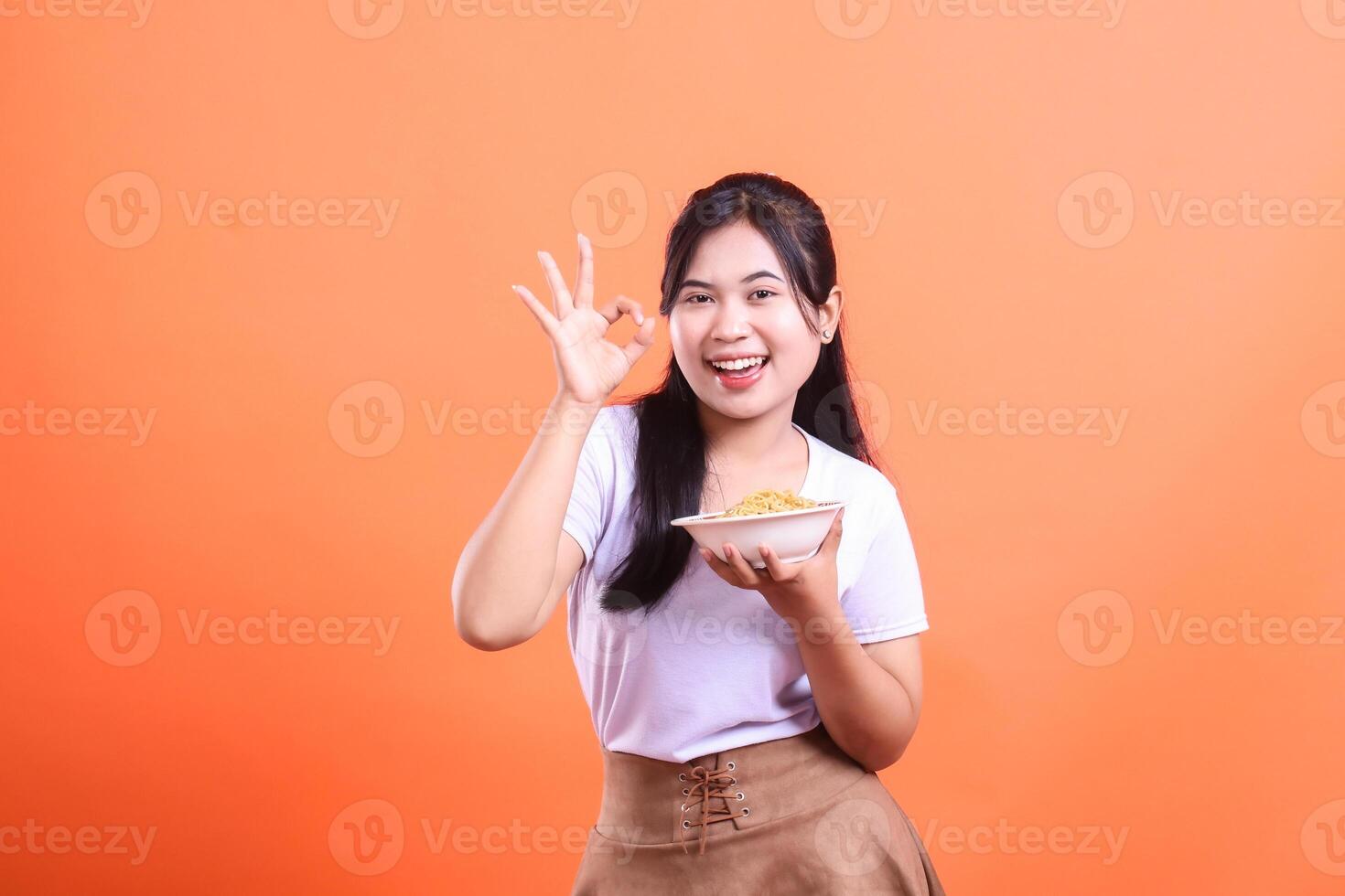 A young and cheerful woman is showing an OK sign while holding a bowl of noodles, it expresses positivity and satisfaction isolated on orange background photo