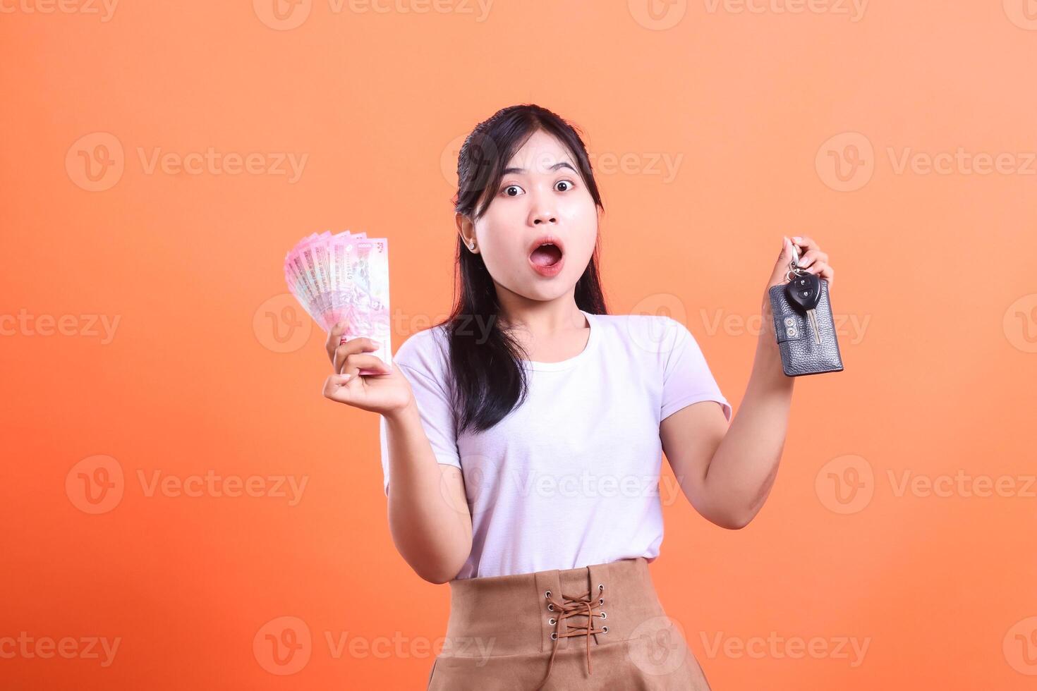 Excited young woman holding money and car key with surprised expression, symbolizing financial success and new beginnings. isolated on orange background photo