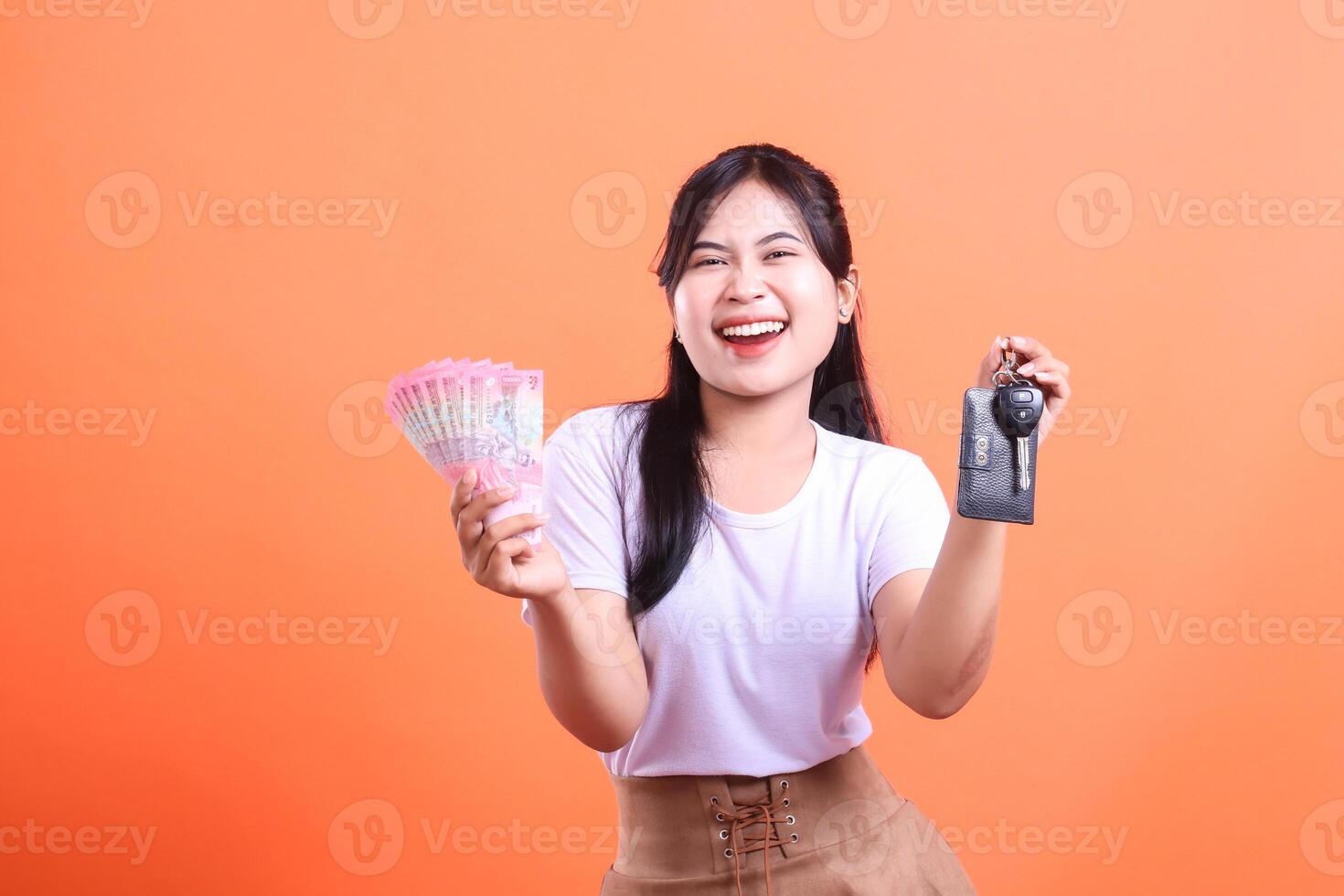Smiling Asian woman holding money and car key, depicting success and financial freedom. isolated on orange background. photo