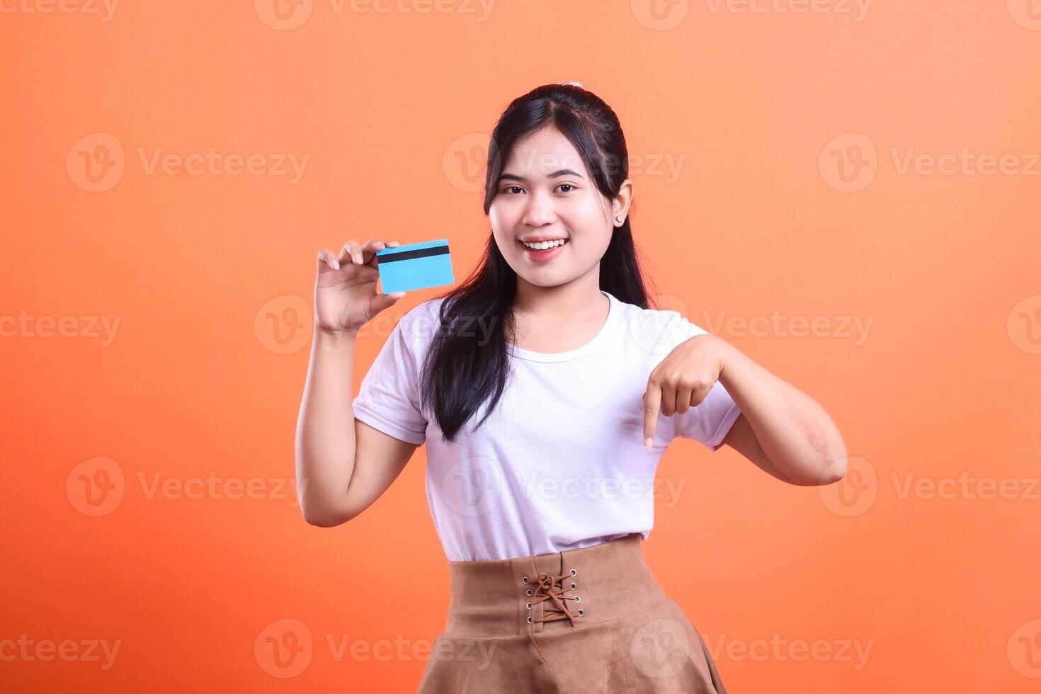 A cheerful young woman holding a blue credit card, smiling and pointing down with her right hand, isolated on orange background photo