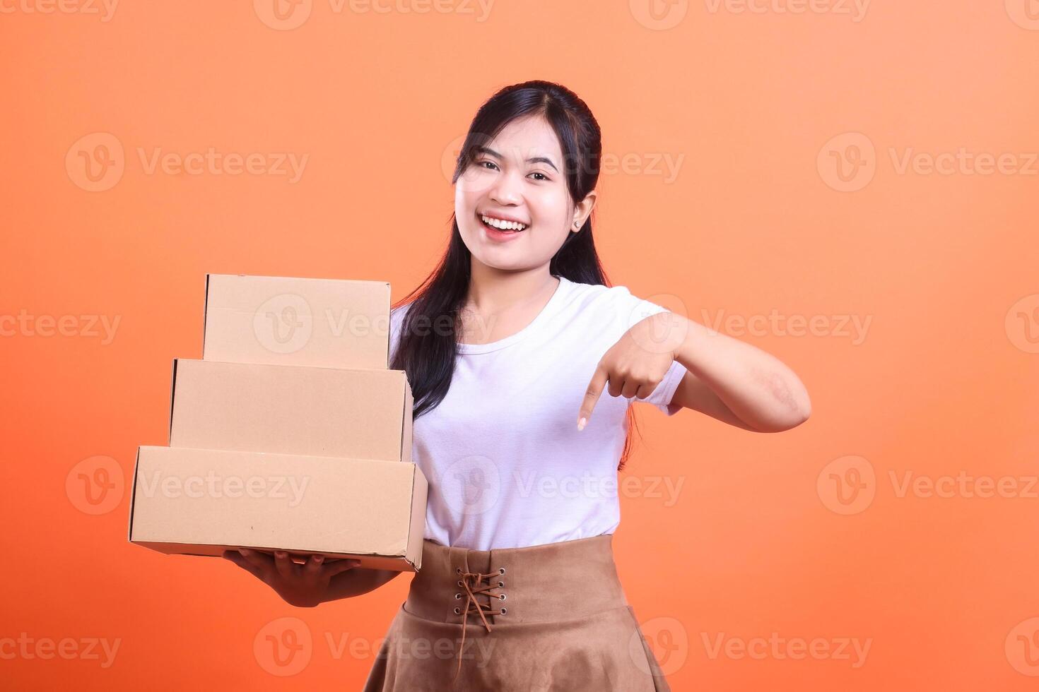 A happy woman holds a stack of cardboard boxes while pointing at them with her finger, she is smiling and cheerful. isolated on orange background photo