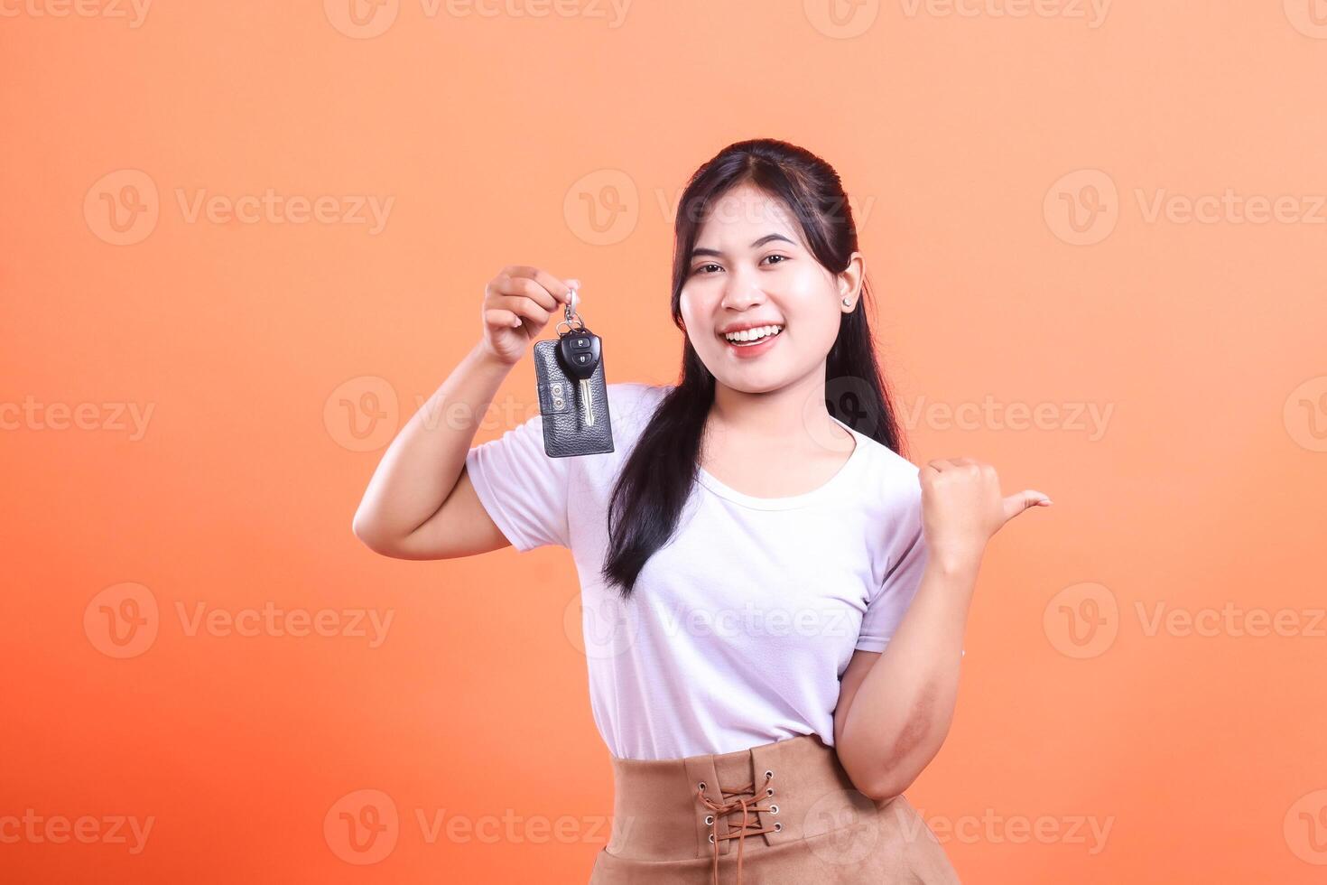 Young woman holding a car key, smiling and showing a thumb on her side, to present something. isolated on orange background photo