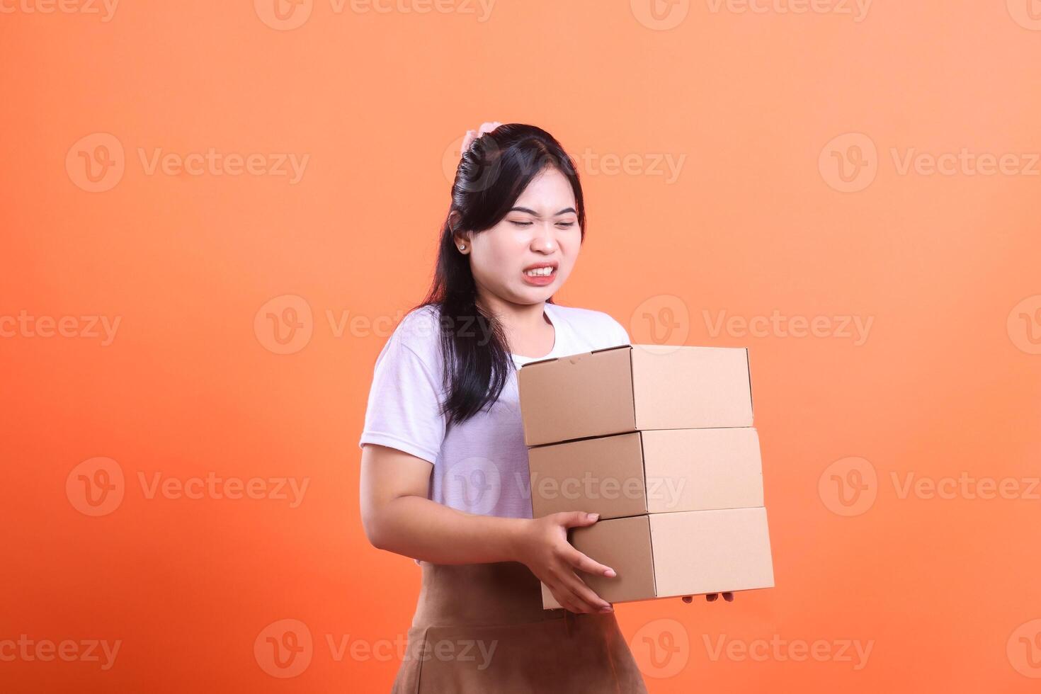 A young woman grimaces while carrying a stack of heavy cardboard boxes with difficulty, expressing physical strain, isolated on orange background photo