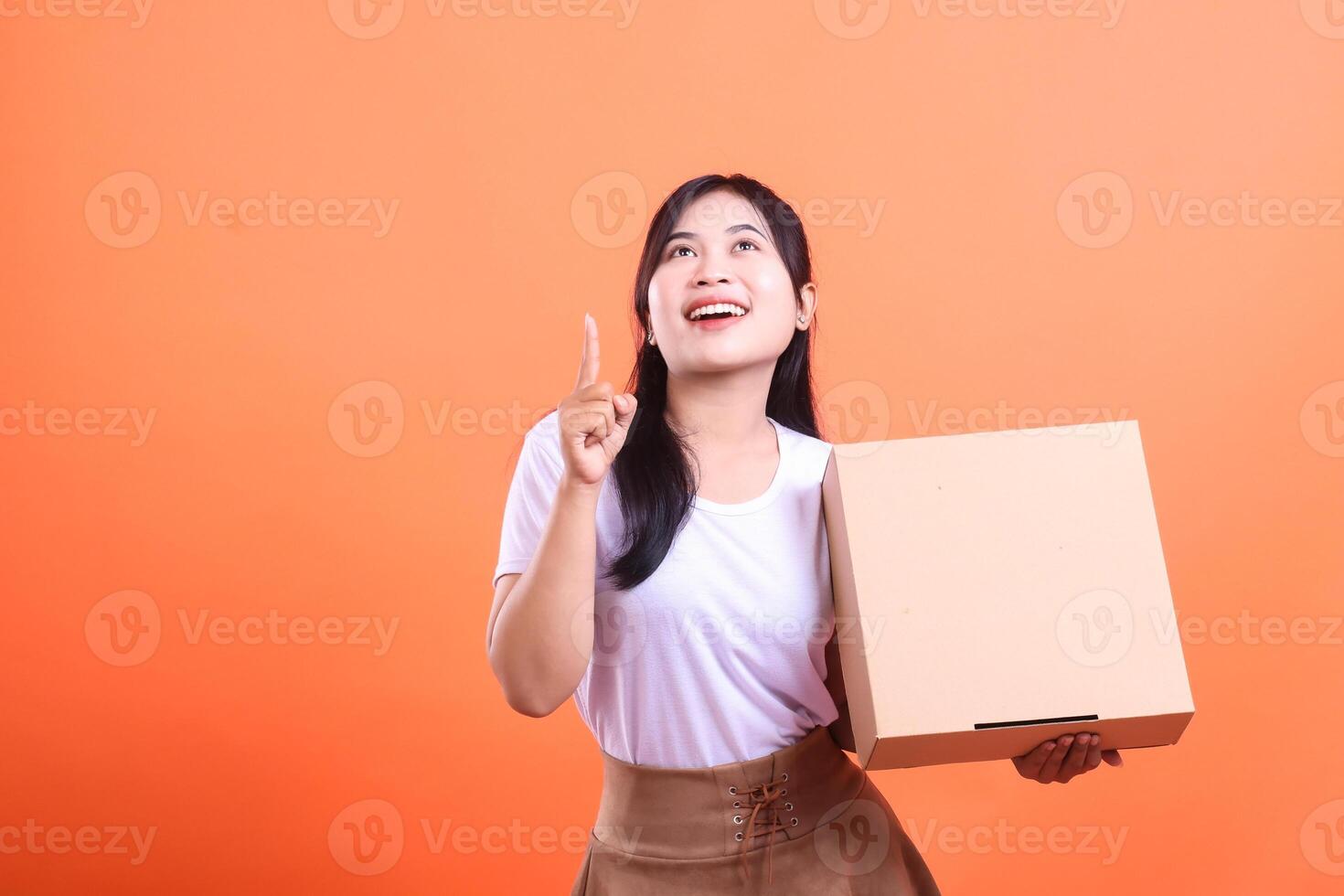 A cheerful woman holds a cardboard box while pointing up with her index finger, suggesting a great idea or a happy purchase isolated on orange background photo