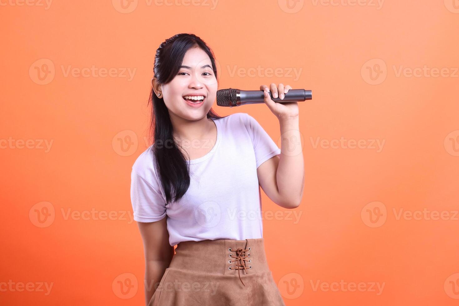 A smiling young woman holding a microphone, ready to sing a song or deliver a speech, captured mid-performance. isolated on orange background photo