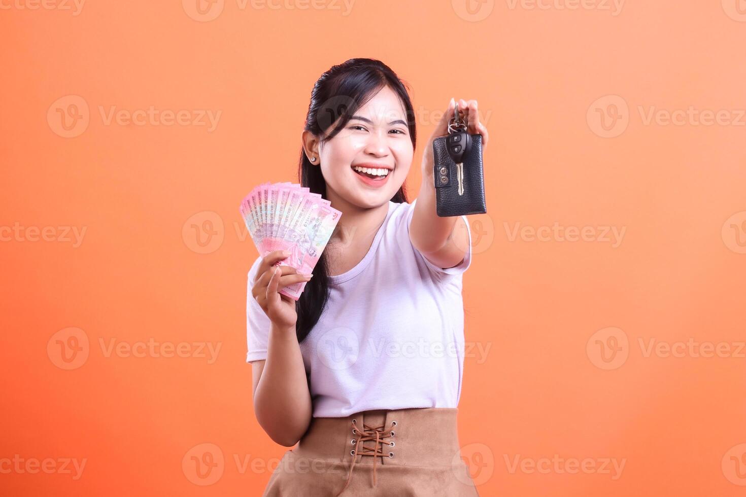 A cheerful Indonesian woman holds a fan of Rupiah and a car key, expressing success and happiness. isolated on orange background. photo