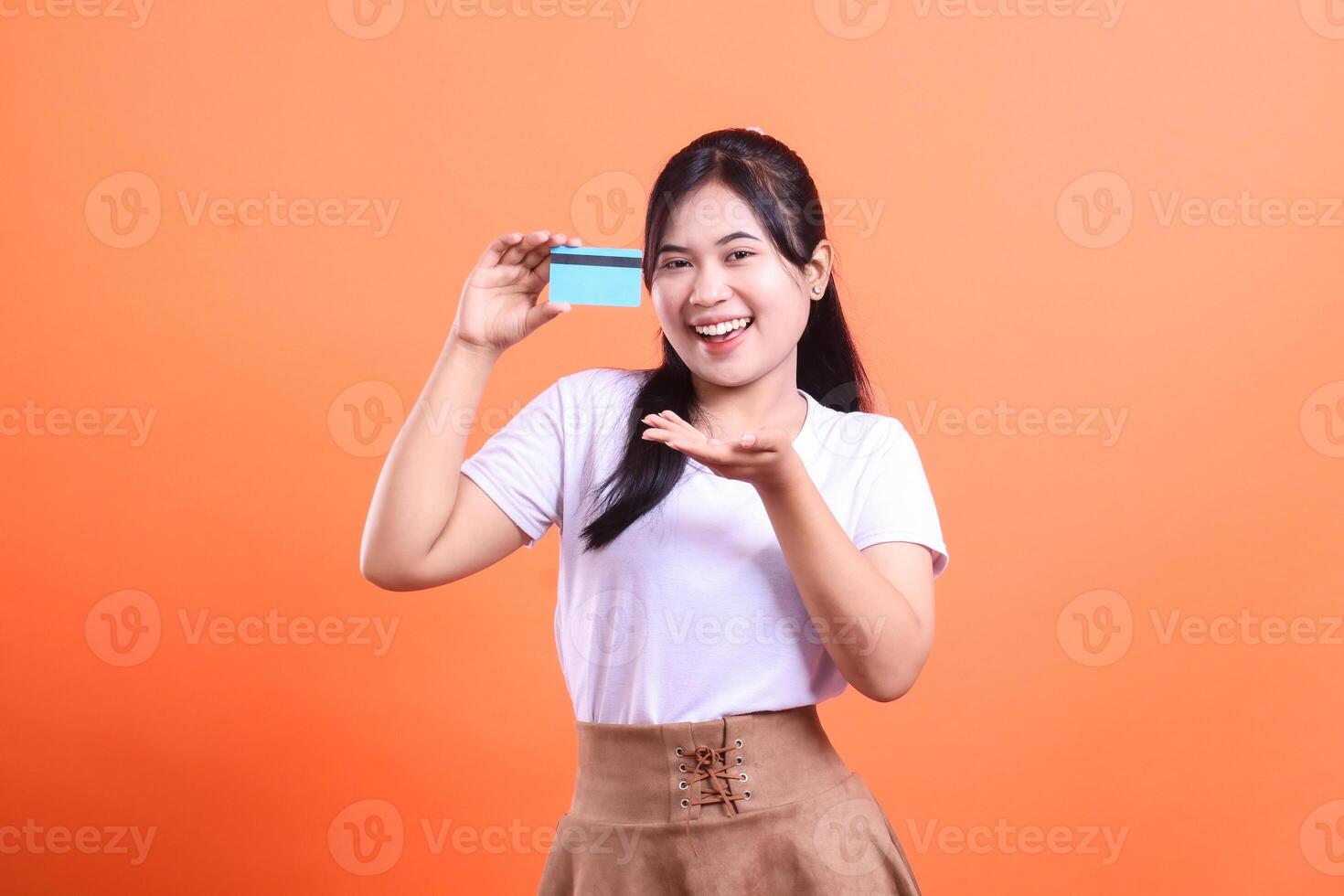 Cheerful young woman holding a credit card and smiling, showcasing the convenience of online transactions and financial access isolated on orange background photo