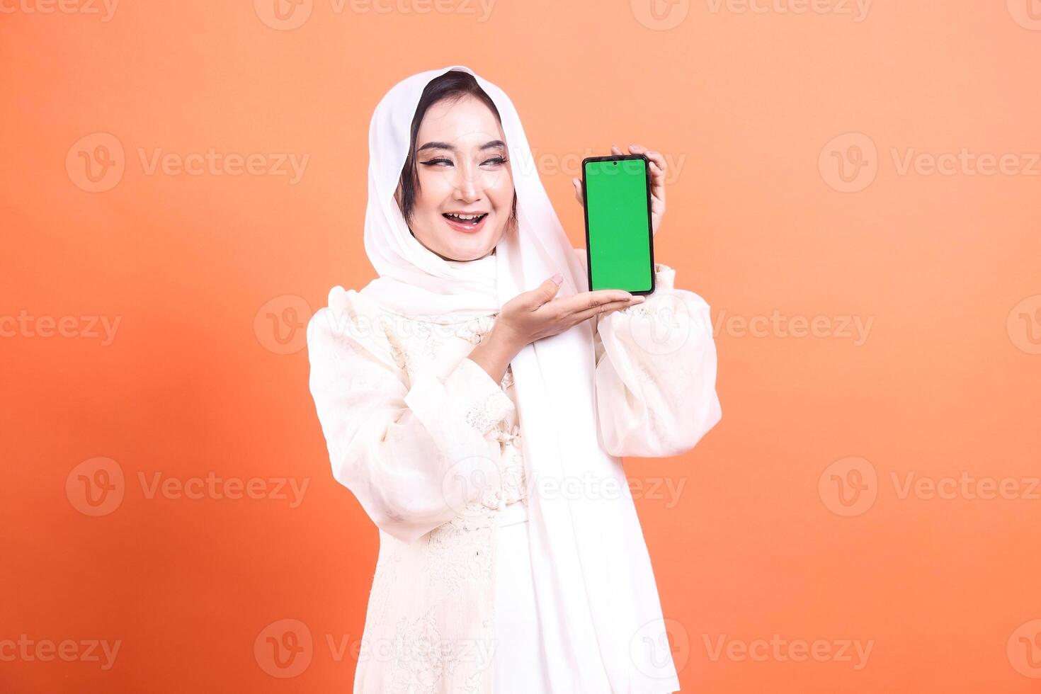 A cheerful Muslim woman in hijab presents a green screen phone with a bright smile, inviting engagement. isolated on a orange background photo