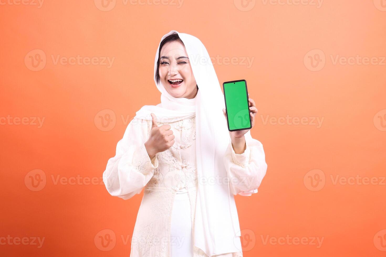 A cheerful Muslim woman in hijab holds up a smartphone with a green screen, giving a thumbs up, and smiling. isolated on a orange background photo