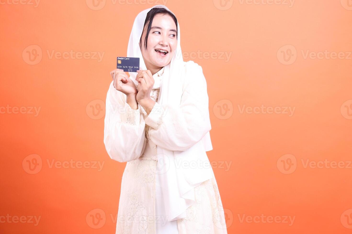 Delighted Muslim Woman Holding Credit Card A bright and cheerful portrait of financial empowerment and modern lifestyle, isolated on a orange background. photo
