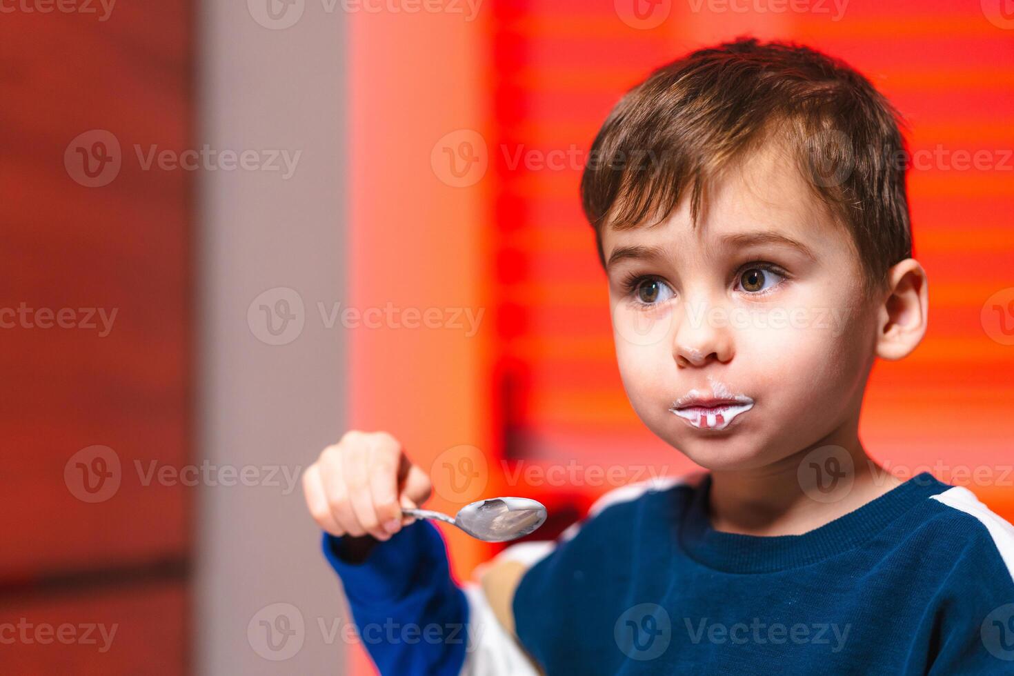 Young boy enjoying ice cream at home. A child with a messy face happily savors a spoonful of ice cream while seated indoors with warm lighting. photo