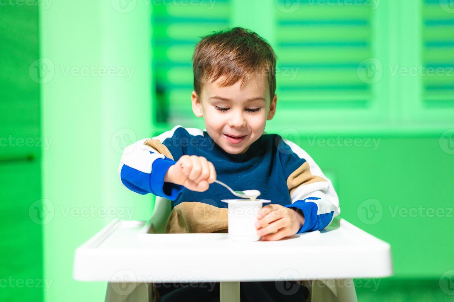 Child enjoying yogurt in a bright room. A young boy sits in a high chair, gleefully eating yogurt with a spoon in a brightly lit green space. photo