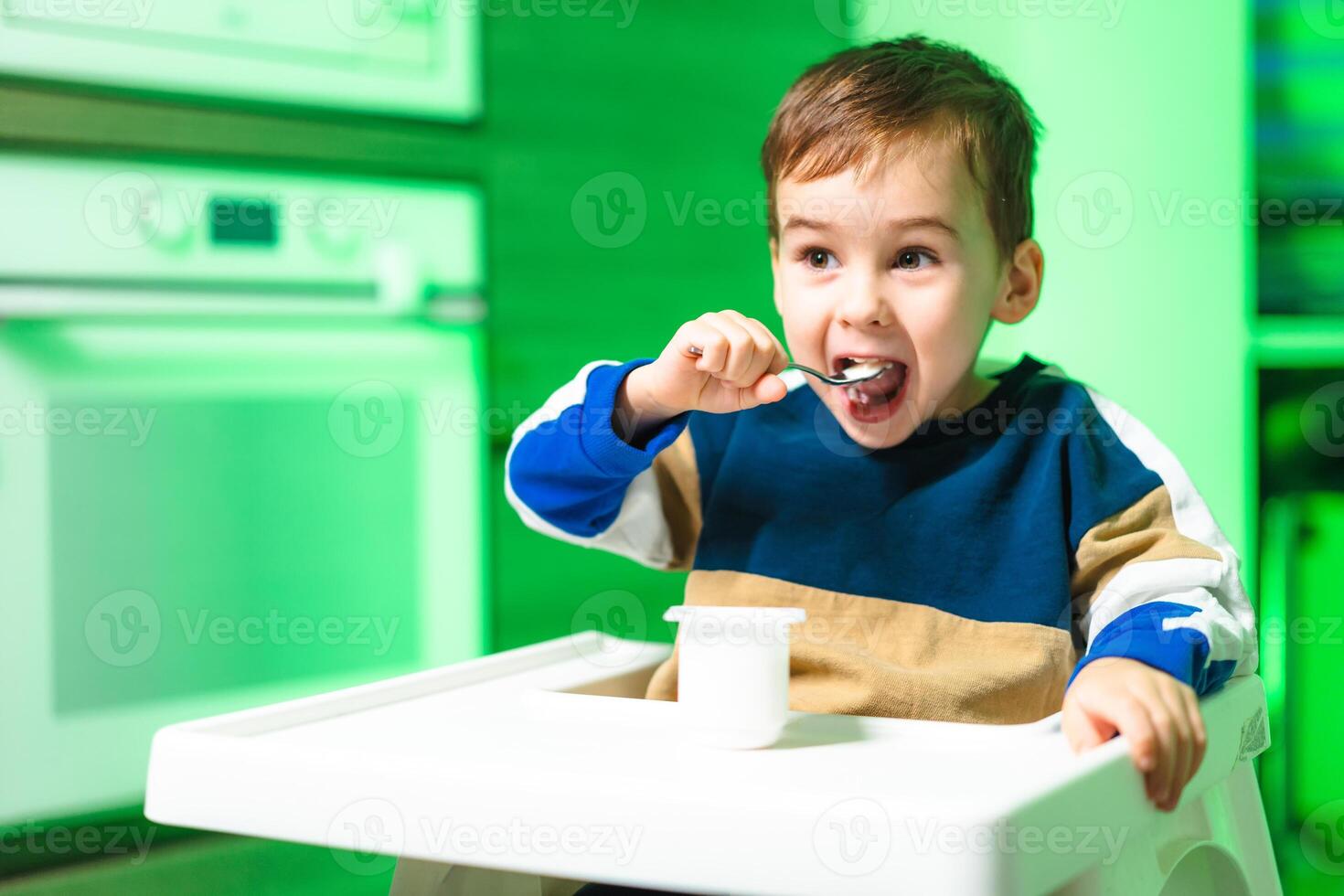 Happy child enjoying yogurt in kitchen. A joyful child sits in a high chair, eating yogurt with a spoon in a brightly lit kitchen during daylight. photo