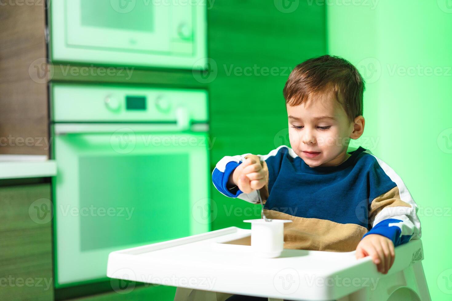 Child enjoying snack in modern kitchen. A young child sits in a high chair, focused on eating yogurt with a spoon in a brightly lit kitchen. photo