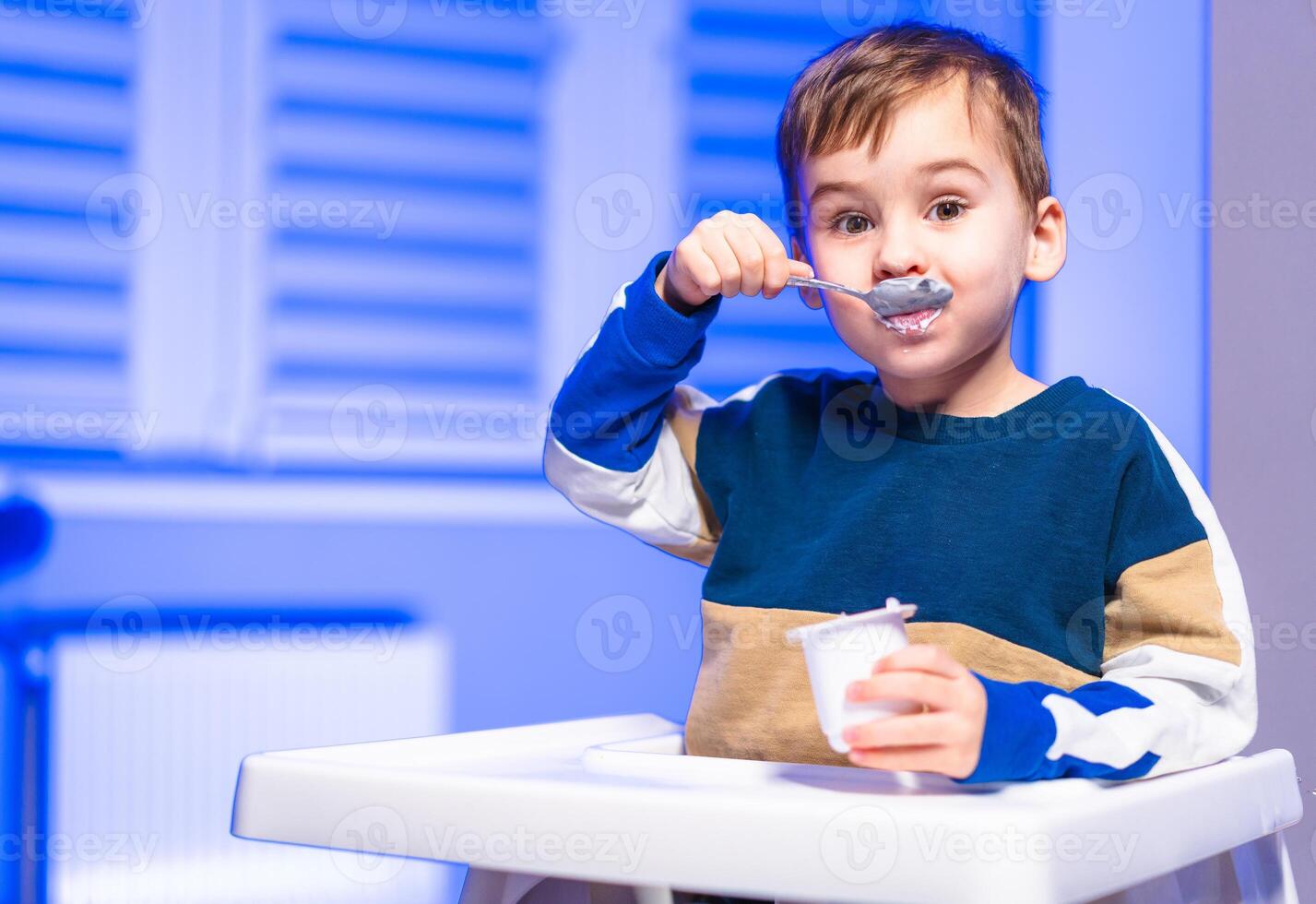 Young child enjoying yogurt at home. A young child is happily eating yogurt while sitting in a high chair, showing joy during snack time. photo