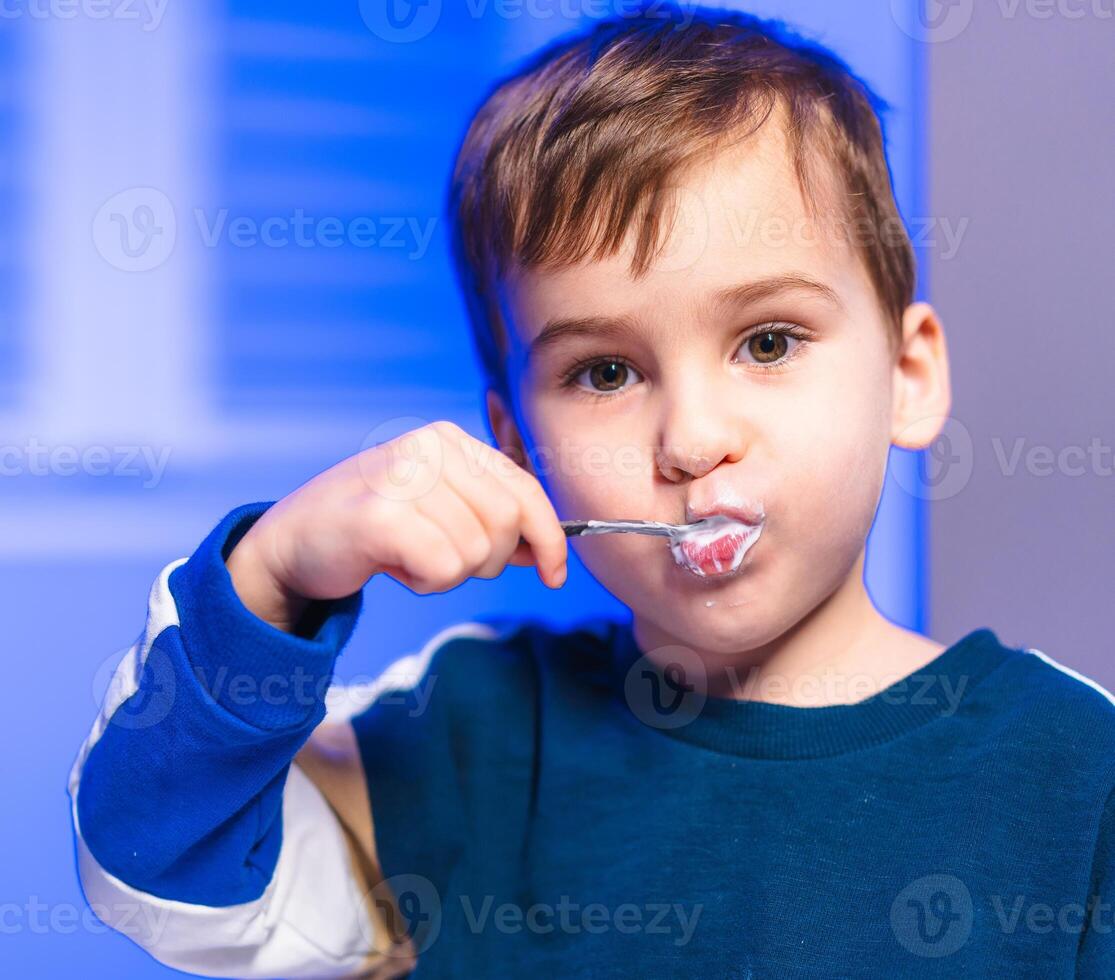 Child eating dessert inside. A young boy happily tastes dessert with a spoon, surrounded by blue lighting at home during the evening. photo