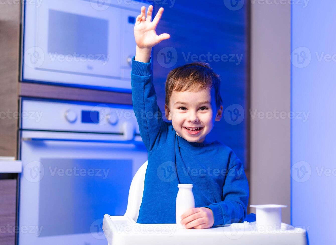 Happy child raising hand in kitchen. A young boy sits in a high chair photo