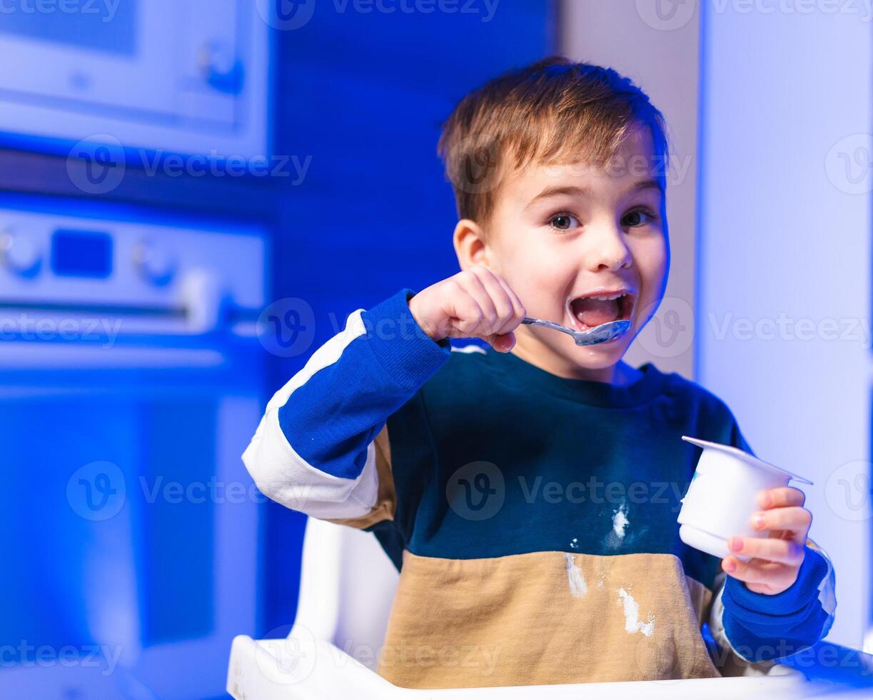 Child enjoys yogurt in kitchen. A cheerful boy enjoys yogurt from a cup, seated in a high chair with a playful expression in a cozy kitchen. photo