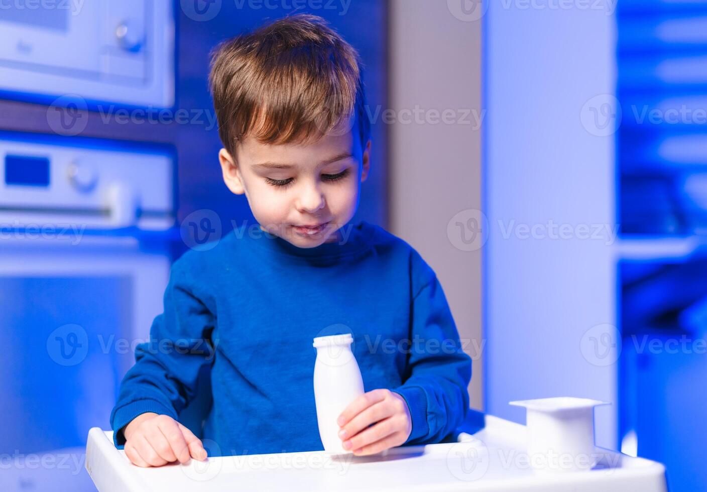 Child with milk bottle in kitchen. A young child in a blue shirt explores a toy milk bottle while standing at a white table in a modern kitchen. photo