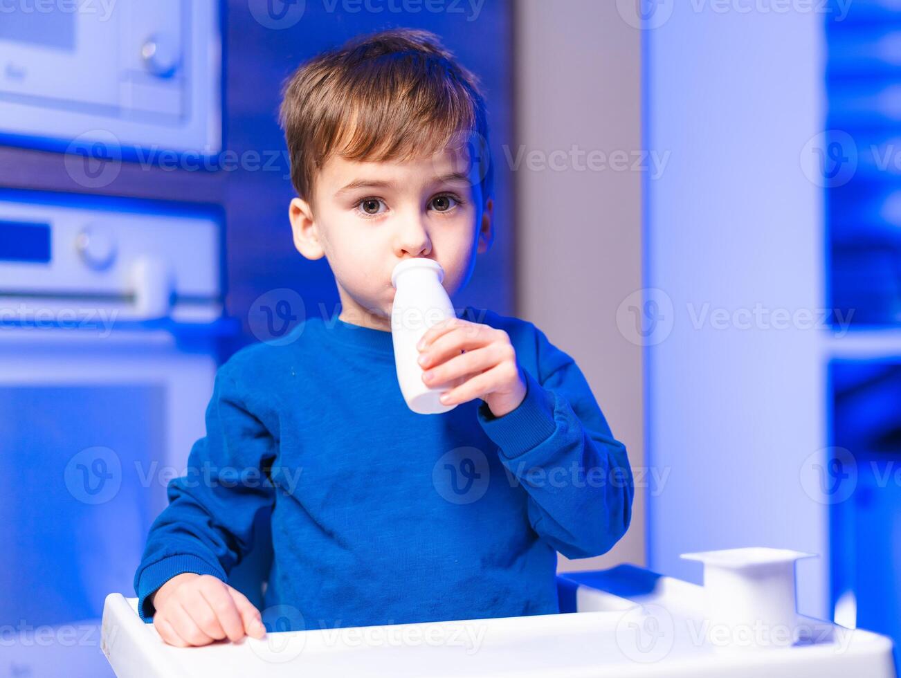 Child with milk in high chair. A young boy is drinking milk while seated in a high chair, surrounded by a modern kitchen setting. photo