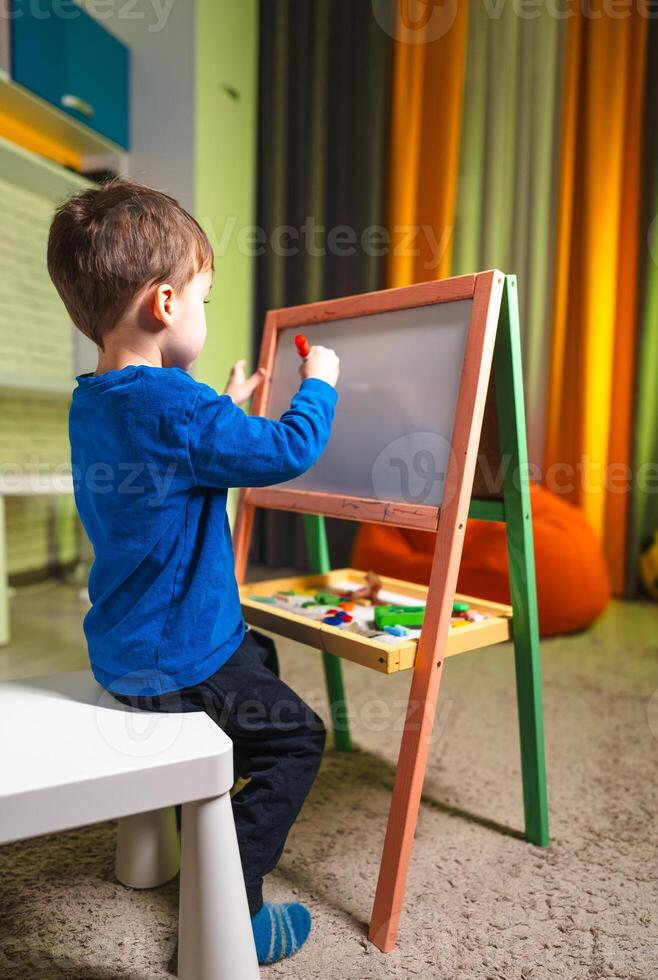 Kid drawing at easel inside. A child focused on creating art at a mini easel in a vibrant indoor space during the day. photo