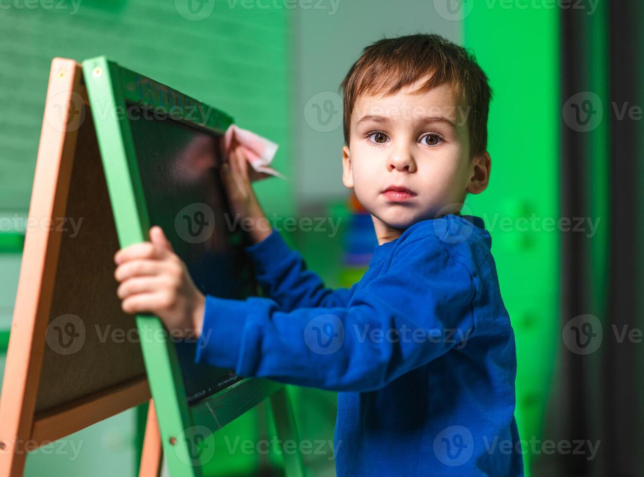 Boy erasing chalkboard in class. A young boy wears a blue shirt as he wipes a chalkboard in a bright, colorful classroom setting. photo