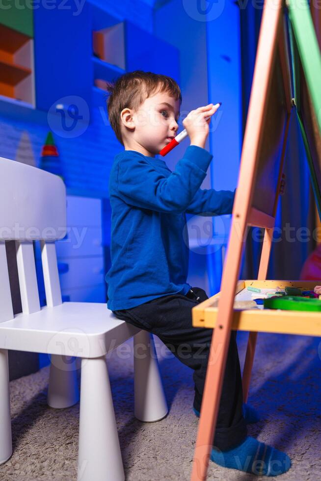 Child loves drawing time. A boy draws at an easel in a bright, colorful room, fully immersed in his creativity. photo