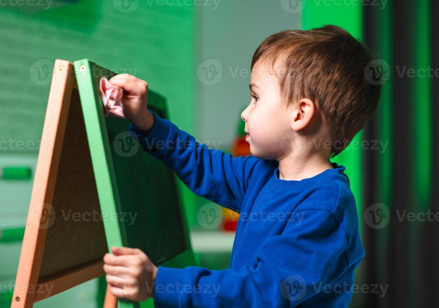 Child erasing chalkboard play. A child in blue cleans a chalkboard while playing indoors, surrounded by vibrant decorations. photo