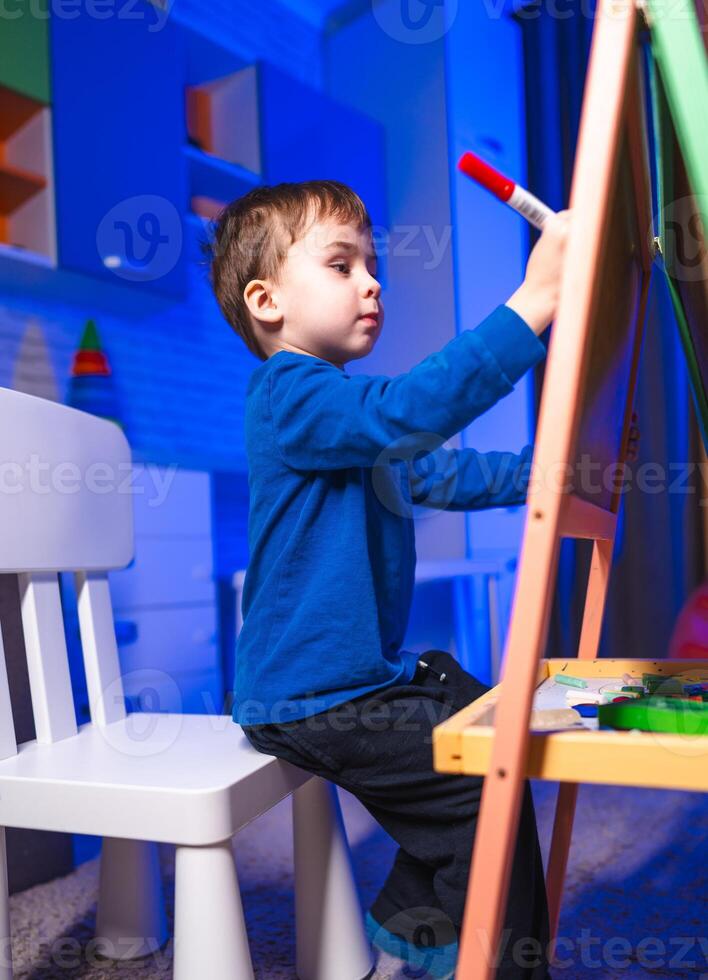 Child happily drawing. A young boy in a blue shirt draws on an easel in a colorful room, focused on his artistic expression at night. photo