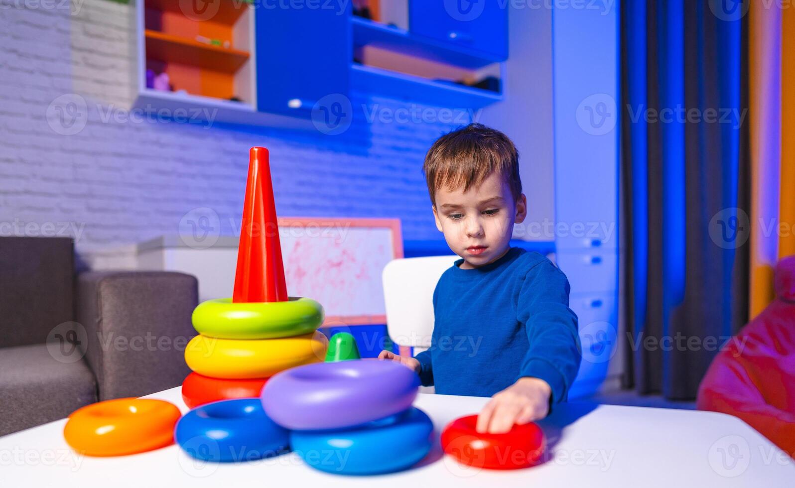 Child playing with stacking toys indoors. A young boy is engaged in stacking colorful toys on a table in a cozy indoor space lit with soft blue hues. photo