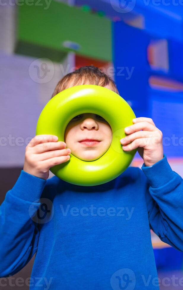 Child plays with green toy ring indoors. A young boy holds a bright green ring in front of his face while enjoying playtime in a colorful indoor environment. photo