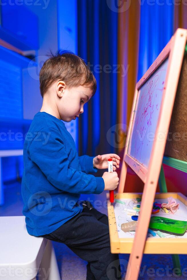Child drawing creatively. A child is focused on drawing with markers at an easel in a brightly lit room, showcasing creativity and concentration. photo