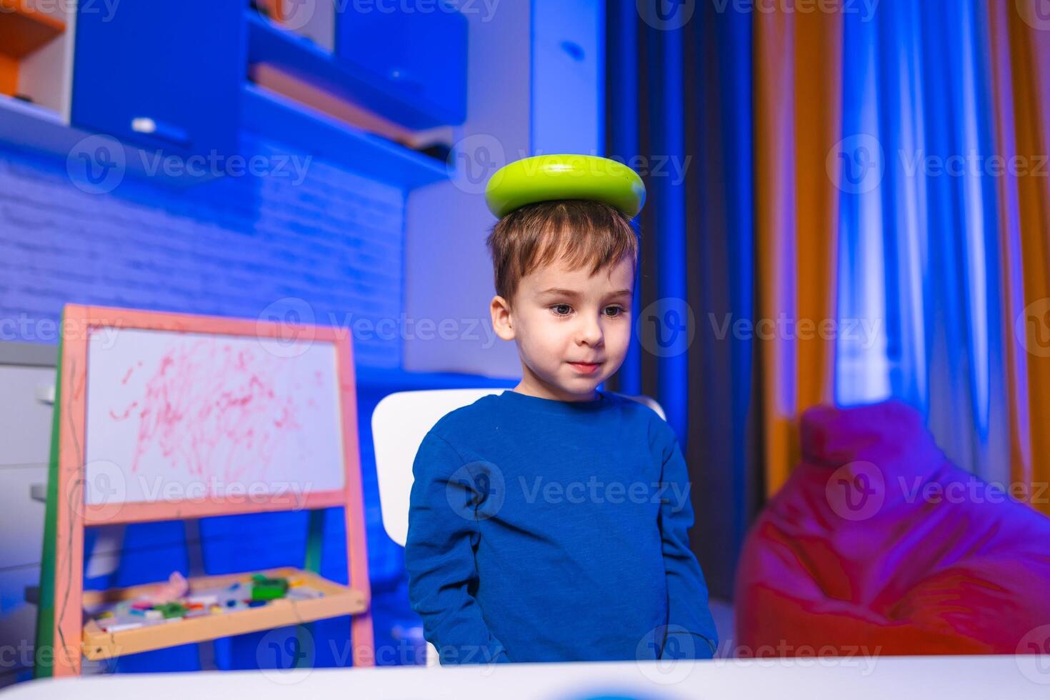 Child balances green item indoors. A young child sits at a table with a green object balanced on their head in a colorful indoor setting. photo