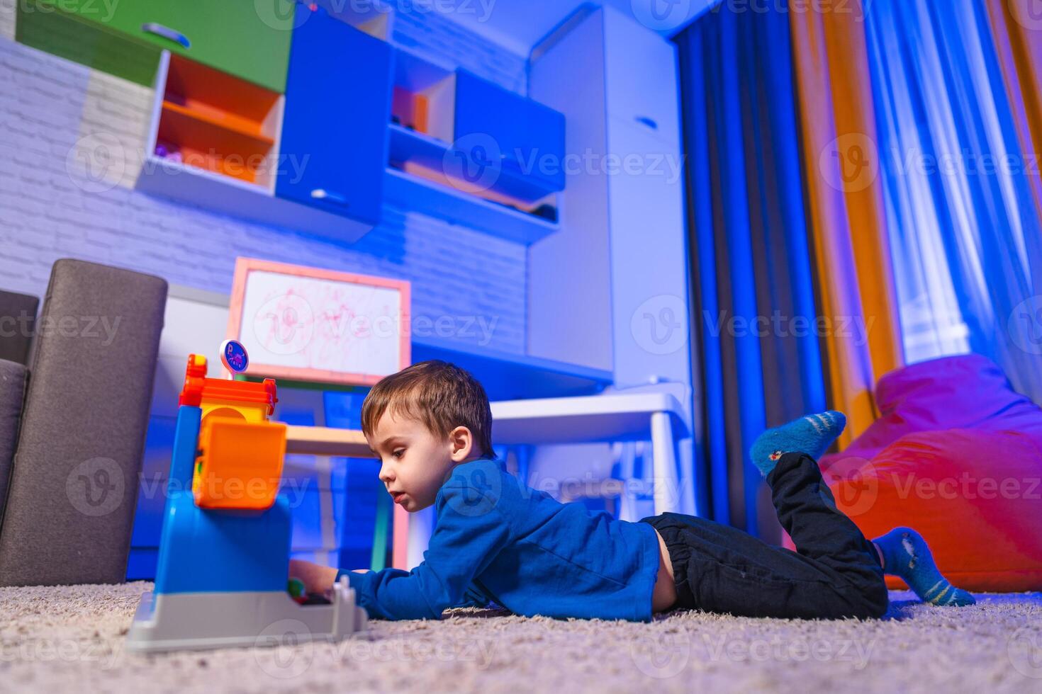 Child plays indoors. A young boy lies on a soft carpet, focused on a colorful toy. The room is brightly lit with blue and orange hues. photo
