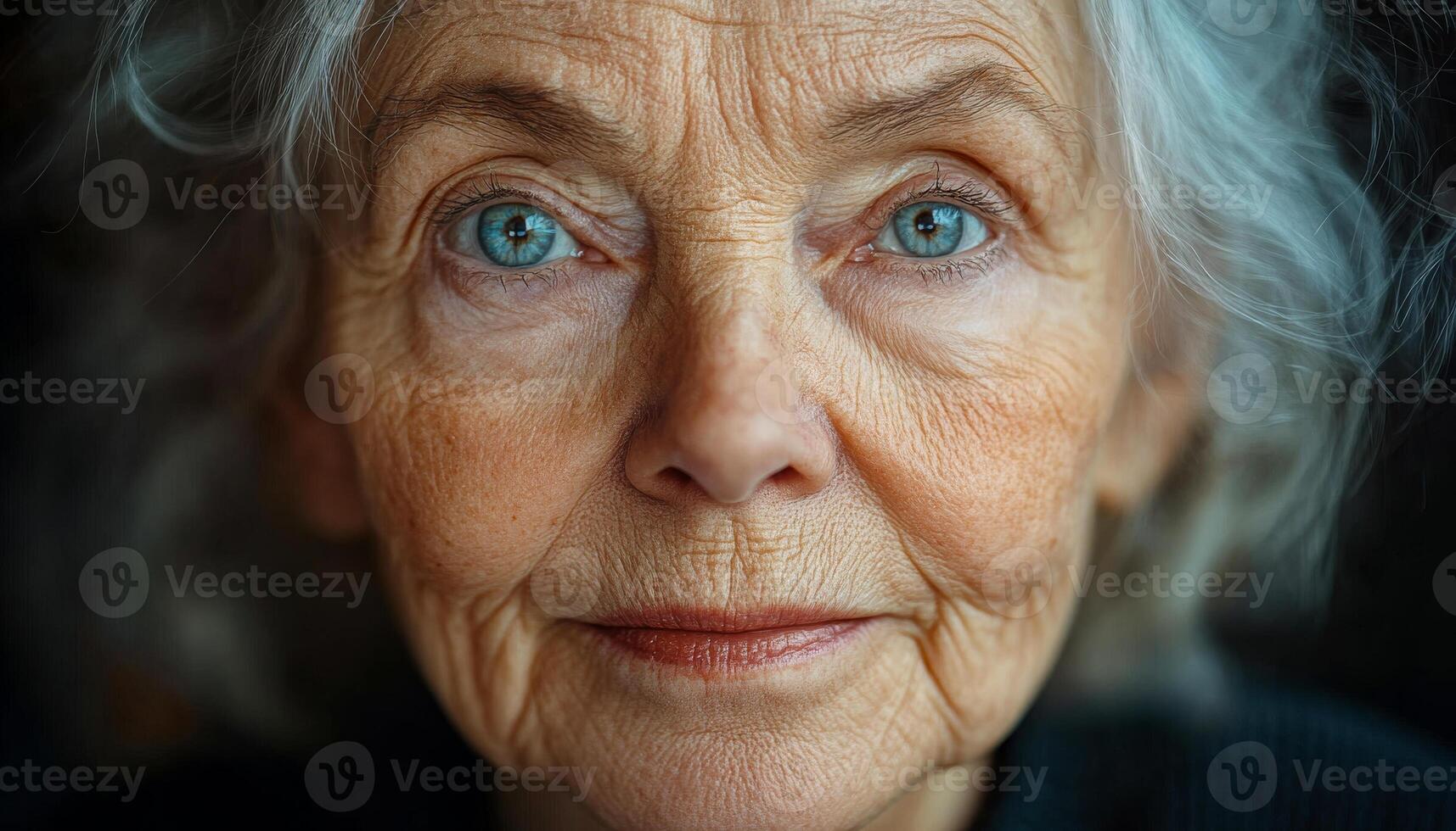 A woman with blue eyes and grey hair is smiling. She has a very wrinkled face and is wearing a black shirt photo