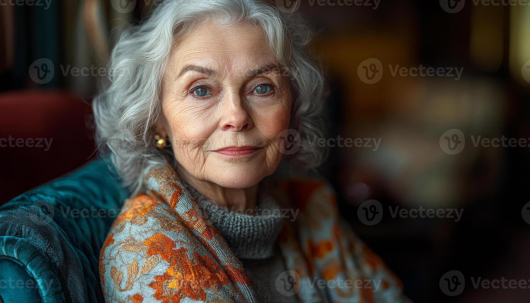 A woman with gray hair and a scarf on her shoulders is sitting on a couch. She has a smile on her face and she is happy photo
