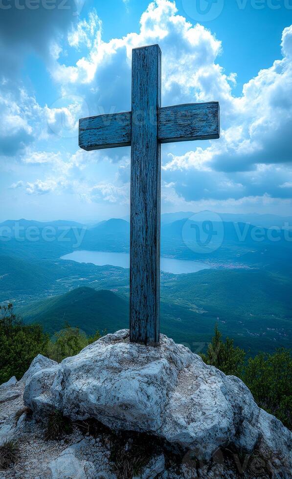 A wooden cross is on a rock in the mountains. The sky is blue and cloudy. The mountains are in the background and there is a lake in the distance photo