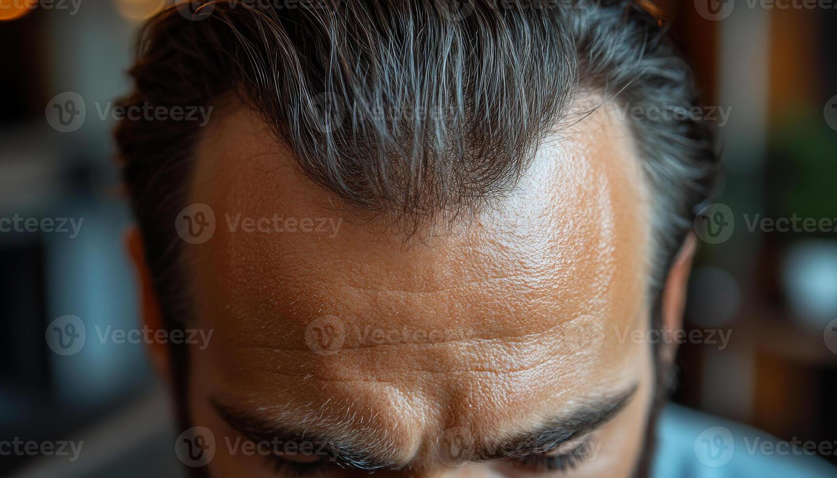 Styled hair man indoors. A man with neatly styled dark hair looks down, showcasing his forehead and hairline in a cozy indoor setting. photo