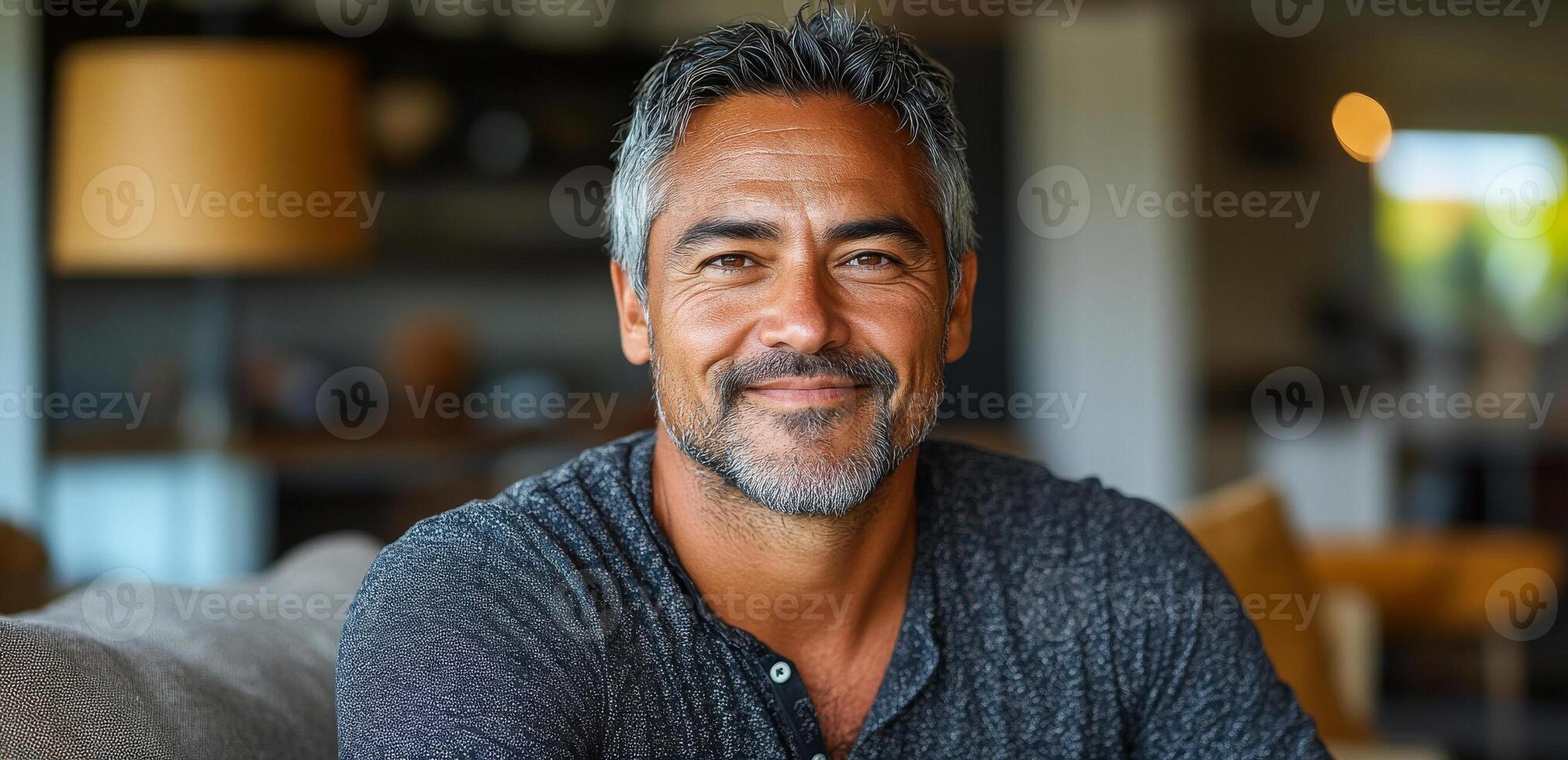 A man with a beard and gray hair is smiling at the camera. He is wearing a black shirt and sitting on a couch photo