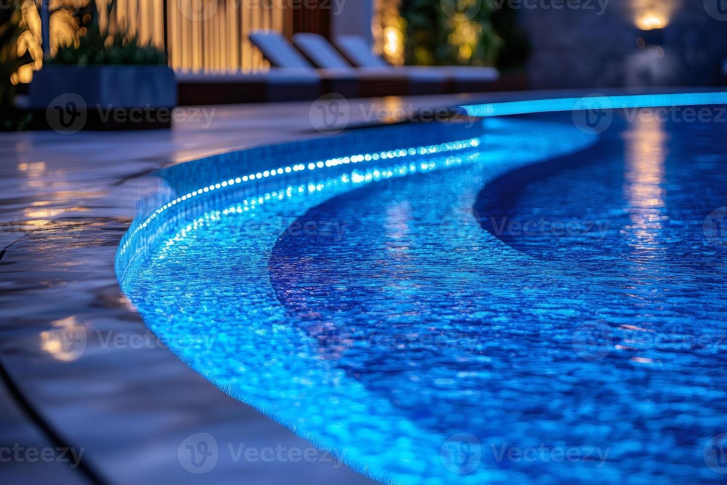 Illuminated luxury resort pool glowing at night with deck chairs in background photo