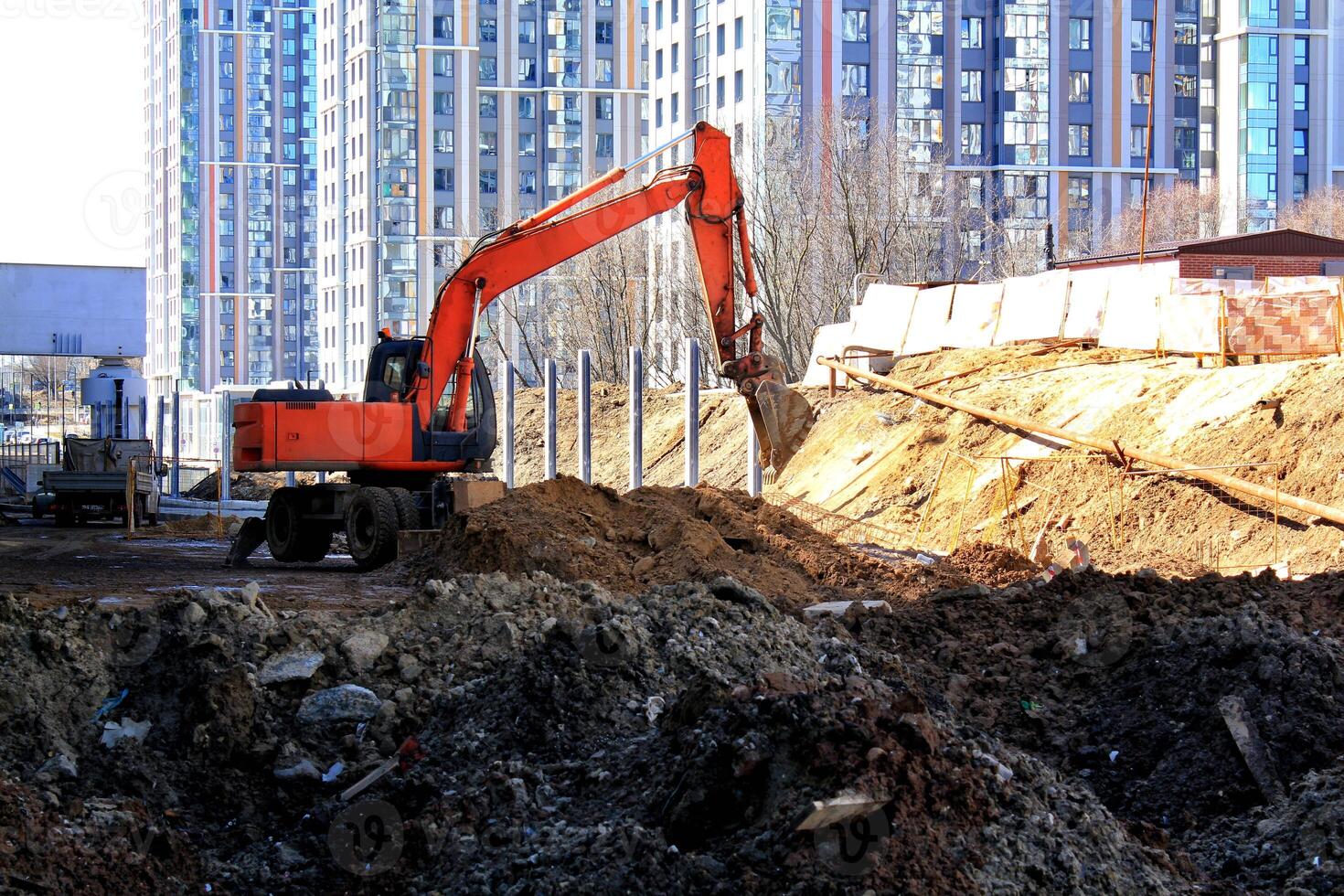 Orange excavator digging at a construction site, with a large building under construction in the background. Construction equipment, urban development, and infrastructure project photo