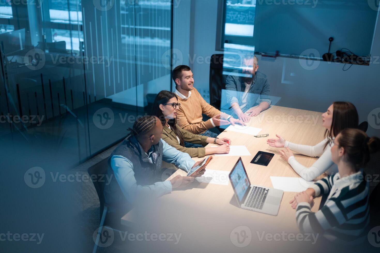 A diverse team of young professionals collaborates in a modern office conference room. Businesspeople discuss strategy, technology, and planning, using laptops to develop an innovative e-commerce photo