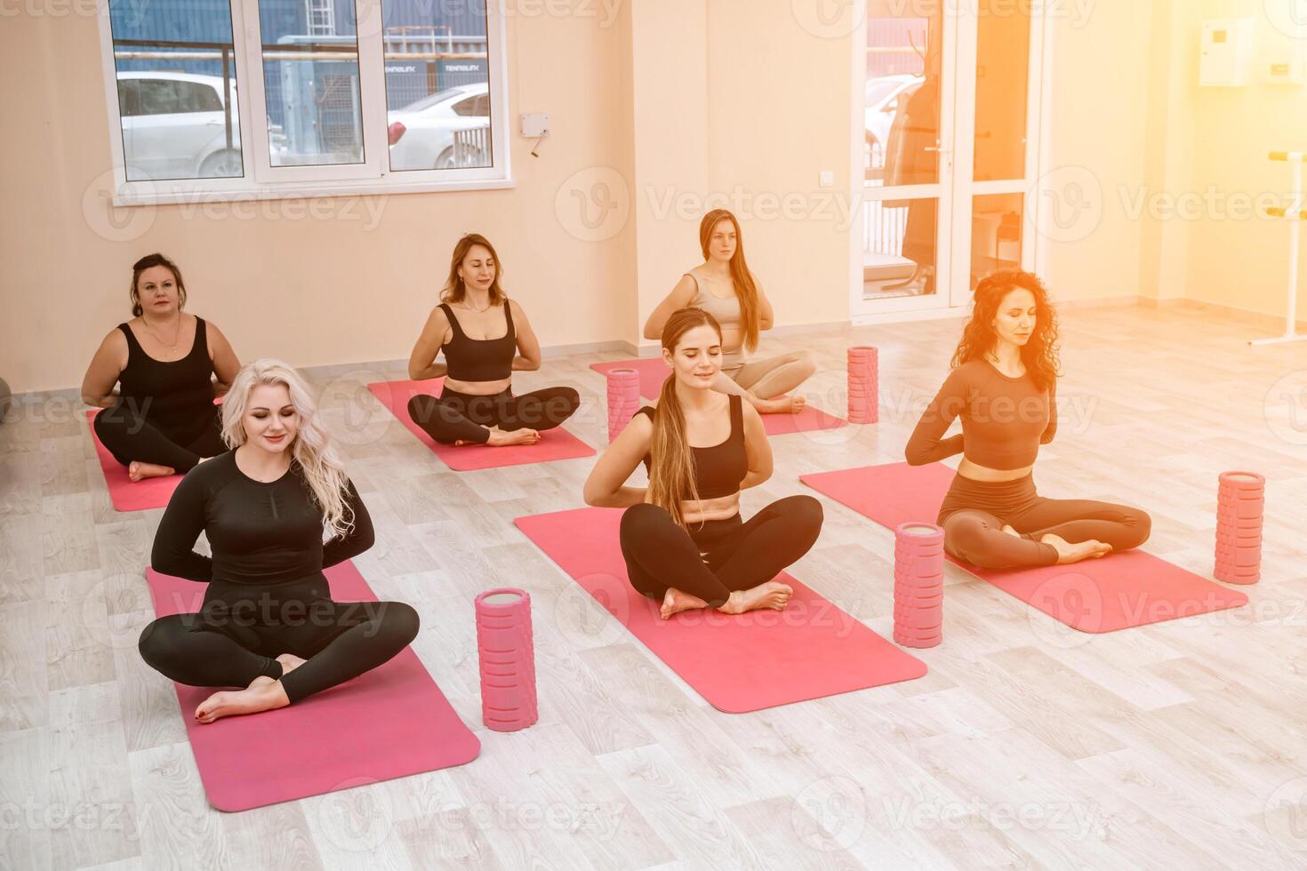 A group of six athletic women doing pilates or yoga on pink mats in front of a window in a beige loft studio interior. Teamwork, good mood and healthy lifestyle concept. photo