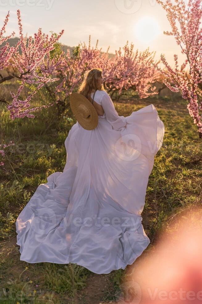Woman blooming peach orchard. Against the backdrop of a picturesque peach orchard, a woman in a long white dress and hat enjoys a peaceful walk in the park, surrounded by the beauty of nature. photo